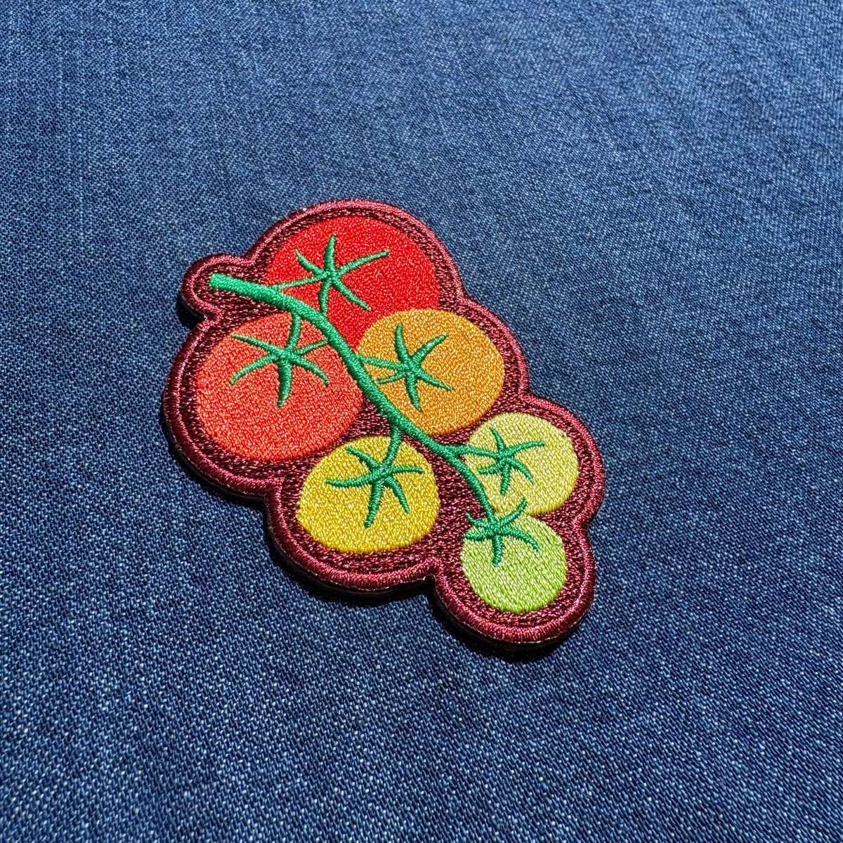 An embroidered patch depicting a cluster of tomatoes on the vine, featuring red, orange, yellow, and green colors, shown on a blue denim fabric background.