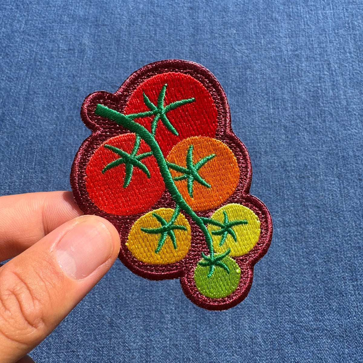 An embroidered patch with red, orange, yellow, and green tomatoes on a vine, held by a hand against a blue denim background.