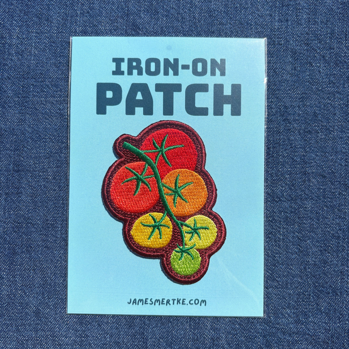 An iron-on patch featuring embroidered tomatoes on a vine, presented on a light blue backing card labeled 'IRON-ON PATCH' with the website 'JAMESMERTKE.COM', set against a dark blue denim background.