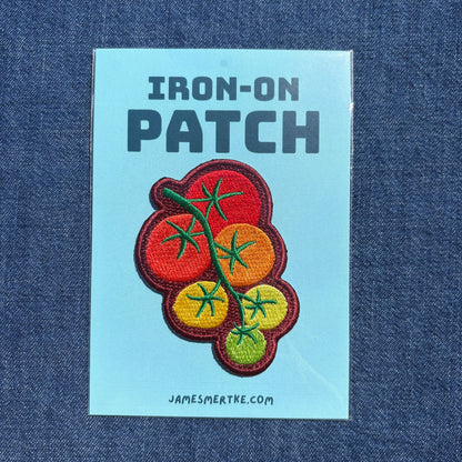 An iron-on patch featuring embroidered tomatoes on a vine, presented on a light blue backing card labeled 'IRON-ON PATCH' with the website 'JAMESMERTKE.COM', set against a dark blue denim background.