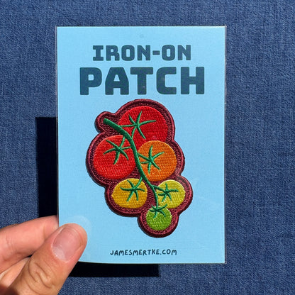 A hand holds a light blue backing card displaying an iron-on patch. The patch features embroidered cherry tomatoes on a vine in red, orange, yellow, and green.