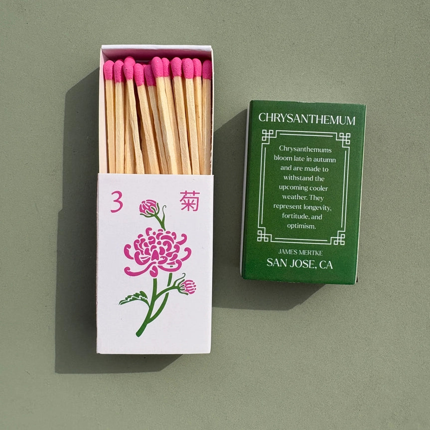 A matchbox is illustrated with a green and pink chrysanthemum to look like a mahjong tile. There is info about the meaning of the plant on the back. Matches with pink heads are inside the box.