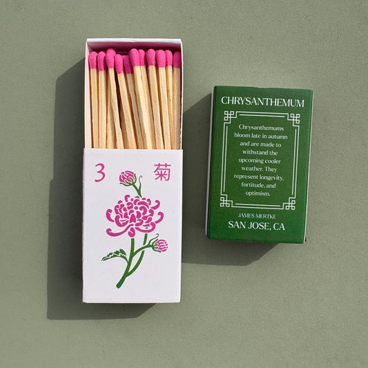 A matchbox is illustrated with a green and pink chrysanthemum to look like a mahjong tile. There is info about the meaning of the plant on the back. Matches with pink heads are inside the box.