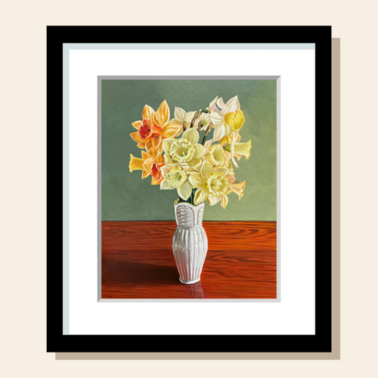 A bouquet of yellow, orange, and white daffodils is gathered in a narrow, white vase. It sits on top of a warm wood grain surface and the wall behind it is a calming green.