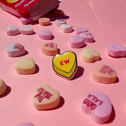 This enamel pin depicts a yellow candy conversation heart with "EW" written on the front in red text.

It is staged with candy hearts and a candy box as props.