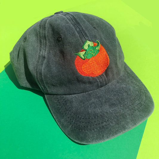 A dark green dad hat is embroidered with a persimmon design.. The fruit os orange and its leaves are green.