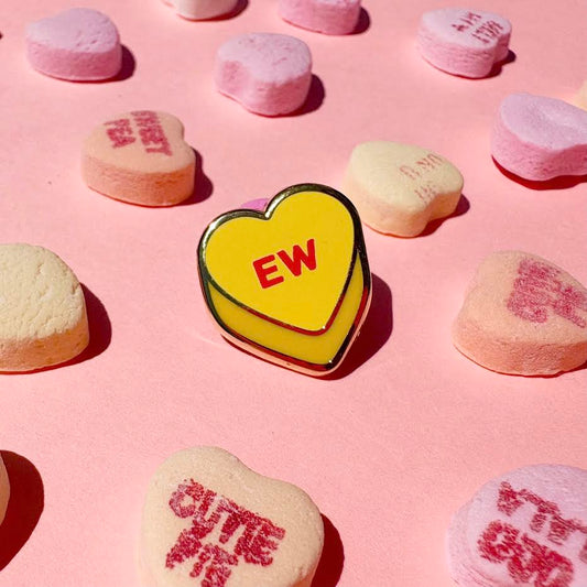 This enamel pin depicts a yellow candy conversation heart with "EW" written on the front in red text.

It is staged with candy hearts as props.