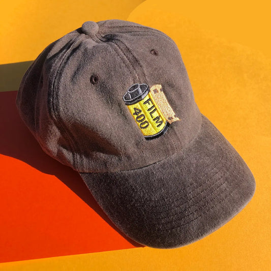 A brown dad hat is embroidered with the illustration of a vintage yellow film canister. The word "FILM" and "400" is one the canister. 