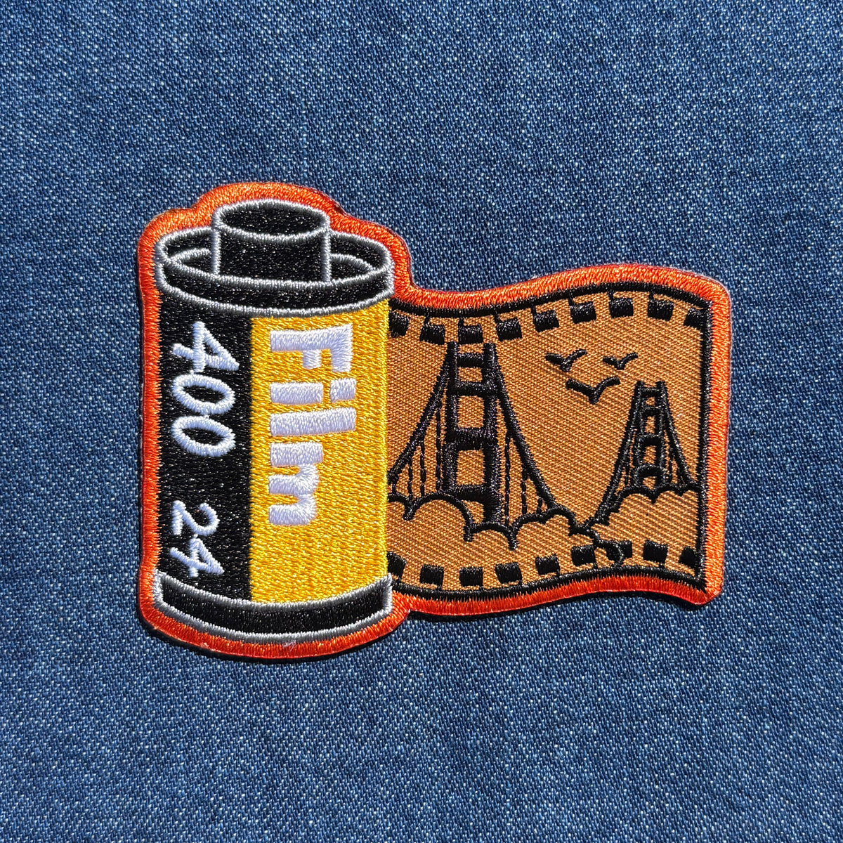 An embroidered patch shaped like a film reel and film strip, featuring a yellow and black film canister with white 'Film 400 24' text and an orange-bordered tan film strip with black bridge and bird outlines, shown on a blue denim background.