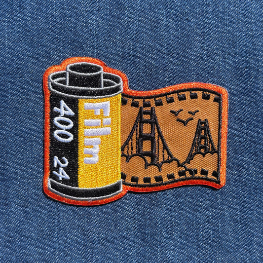 An embroidered patch shaped like a film reel and film strip, featuring a yellow and black film canister with white 'Film 400 24' text and an orange-bordered tan film strip with black bridge and bird outlines, shown on a blue denim background.