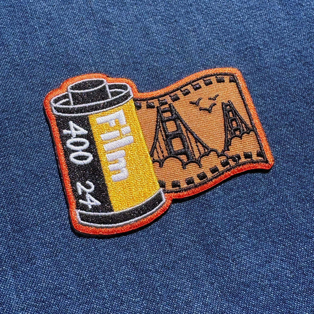 An embroidered patch shaped like a film reel and exposed film strip, featuring a Golden Gate Bridge design, placed on a blue denim fabric background.