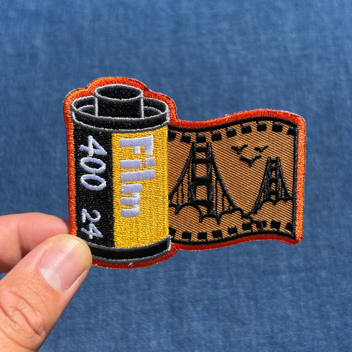 An embroidered patch shaped like a film roll with a partially unrolled strip showing the Golden Gate Bridge, held by a hand against a blue denim background.
