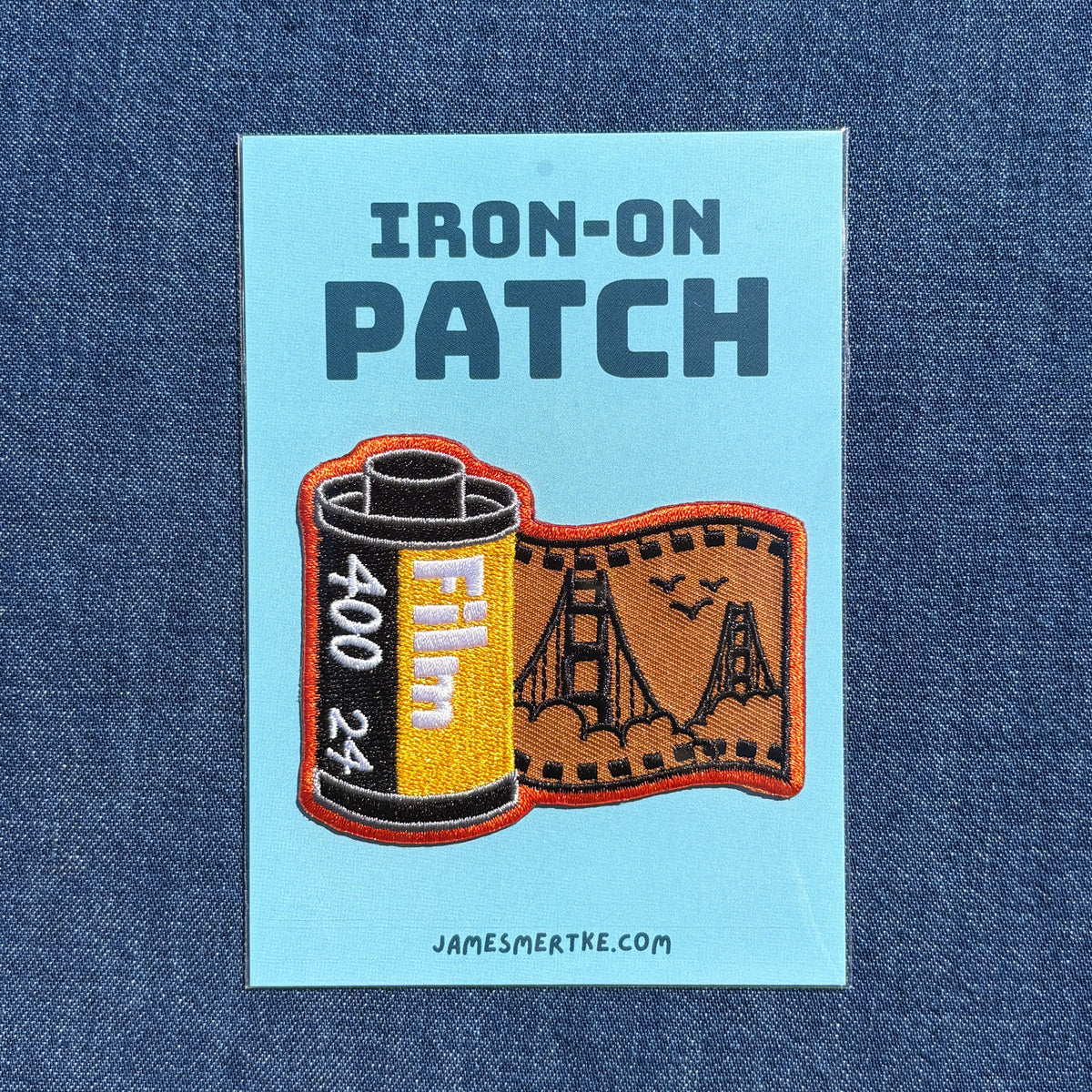 An embroidered patch on a light blue backing card, displayed flat against a blue denim fabric background. The patch features the text 'IRON-ON PATCH' above an embroidered film can and film strip design with the Golden Gate Bridge.