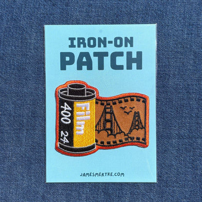 An embroidered patch on a light blue backing card, displayed flat against a blue denim fabric background. The patch features the text 'IRON-ON PATCH' above an embroidered film can and film strip design with the Golden Gate Bridge.