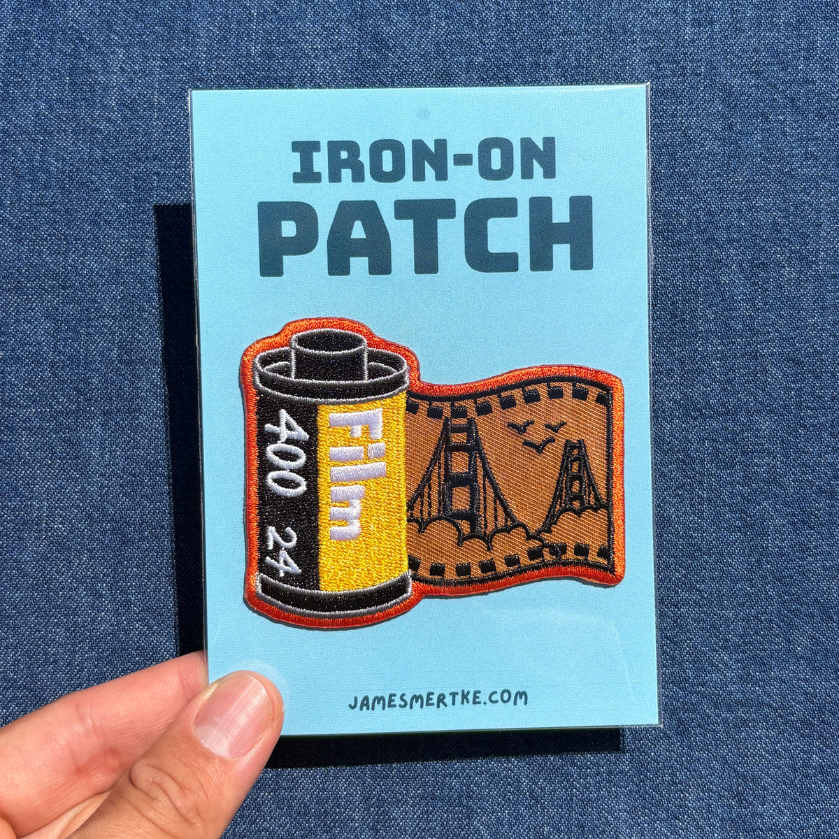 A hand holds a light blue backing card for an iron-on patch against a dark blue denim background. The card reads 'IRON-ON PATCH' and shows a patch design combining a film reel with a Golden Gate Bridge scene.