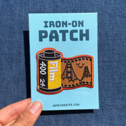 A hand holds a light blue backing card for an iron-on patch against a dark blue denim background. The card reads 'IRON-ON PATCH' and shows a patch design combining a film reel with a Golden Gate Bridge scene.