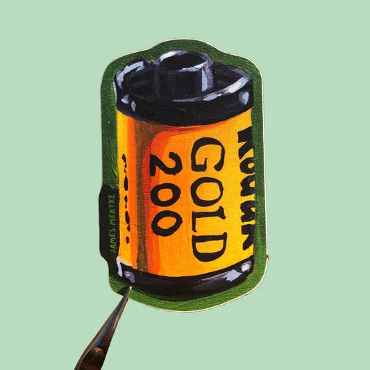 This sticker depicts a painting of an analog film roll. It has a yellow base with the phrase "GOLD 400" written on its side. The background is green.
