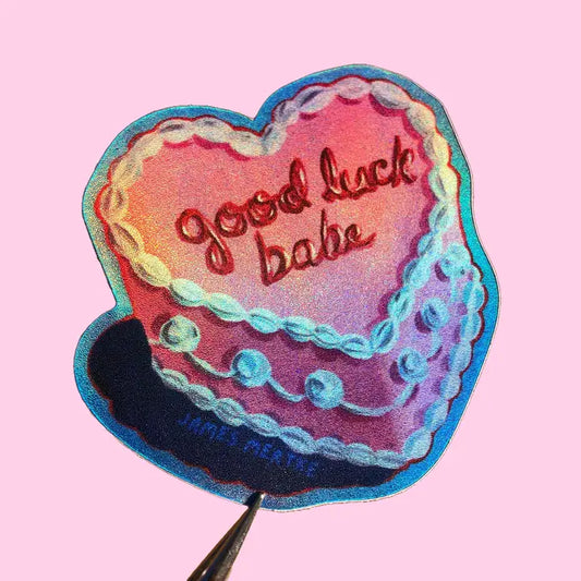 This sticker depicts a painting of a heart shaped cake with pink frosting and white piping details. "good luck babe" is written in cursive on top of the cake in red frosting.  The sticker has a shiny and holographic surface which gives it a special quality.