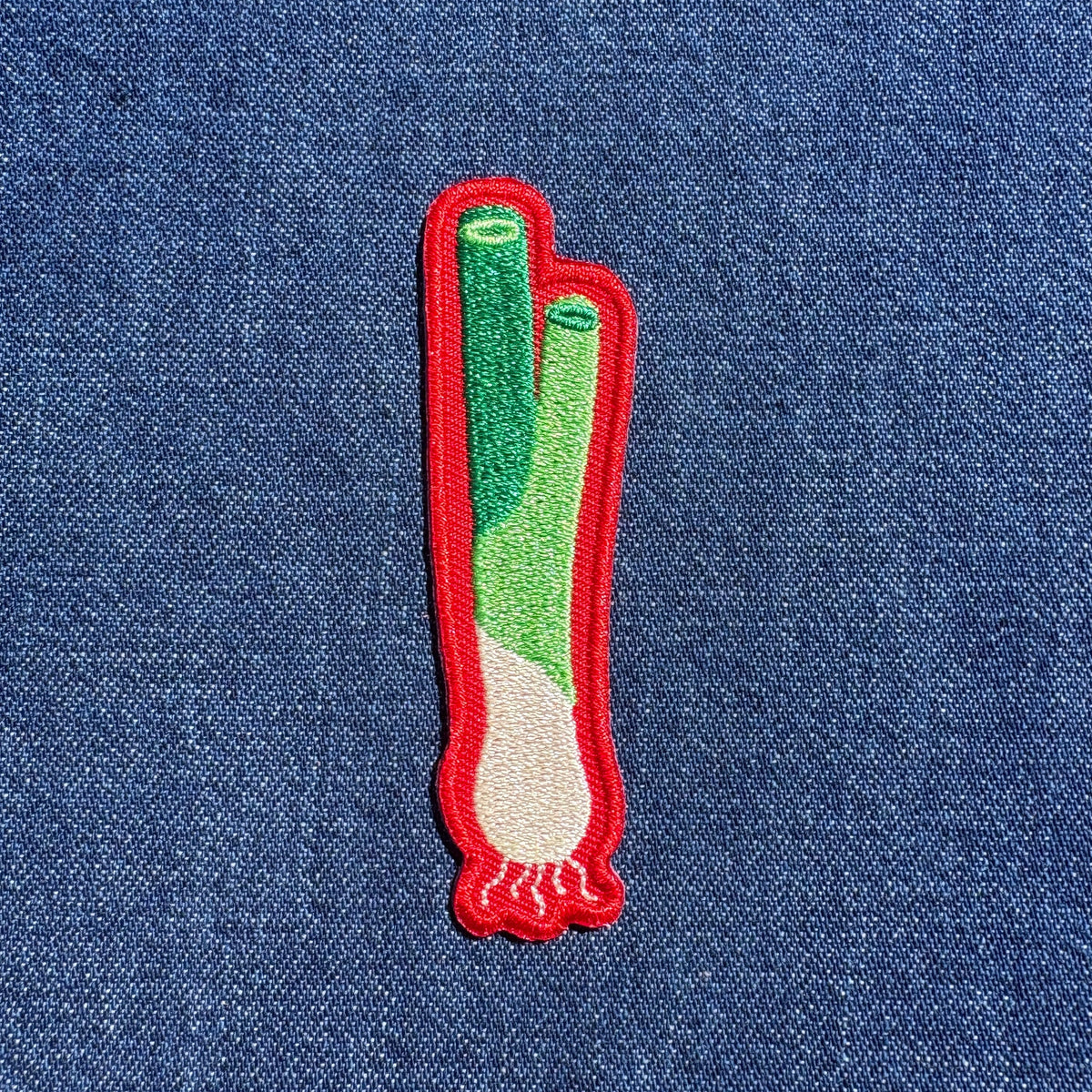 A small embroidered patch shaped like a green onion, featuring green and white stitched details with a red border, placed on a blue denim background.