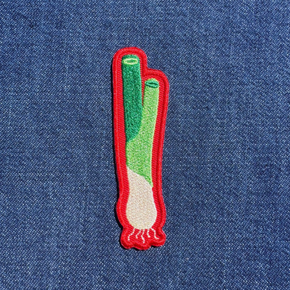 A small embroidered patch shaped like a green onion, featuring green and white stitched details with a red border, placed on a blue denim background.
