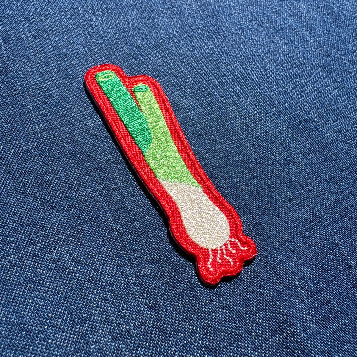 An embroidered patch shaped like a scallion, featuring green and white stitching with a red border, placed on a blue denim fabric background.