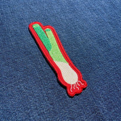 An embroidered patch shaped like a scallion, featuring green and white stitching with a red border, placed on a blue denim fabric background.