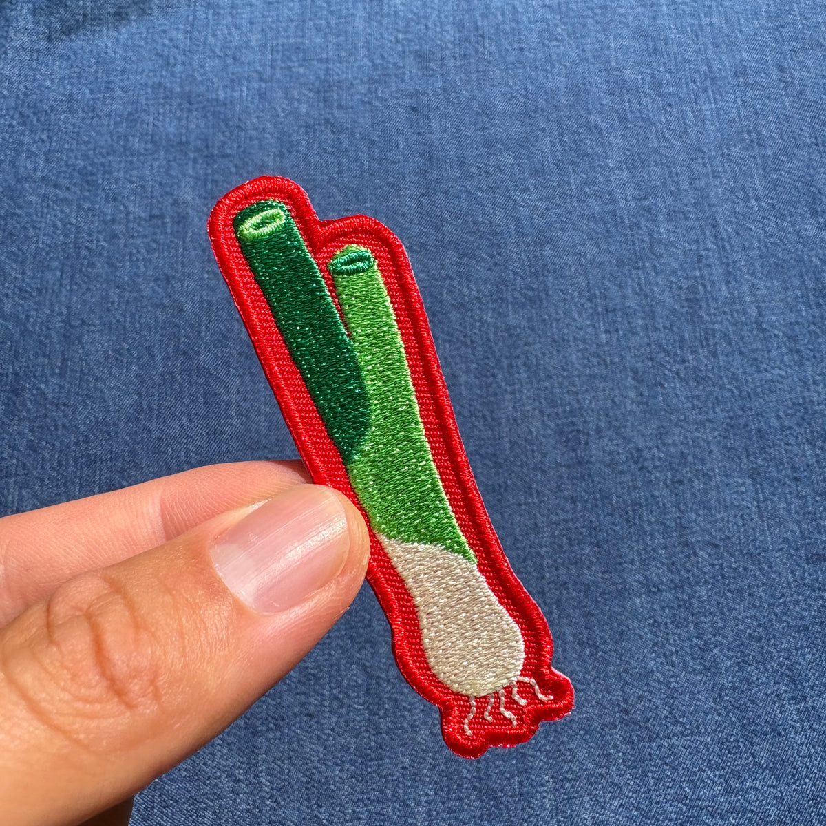 A small embroidered patch shaped like a leek or scallion, held between fingers against a blue denim-like fabric background. The patch has a red border, green upper sections, a white lower bulbous section, and small roots.