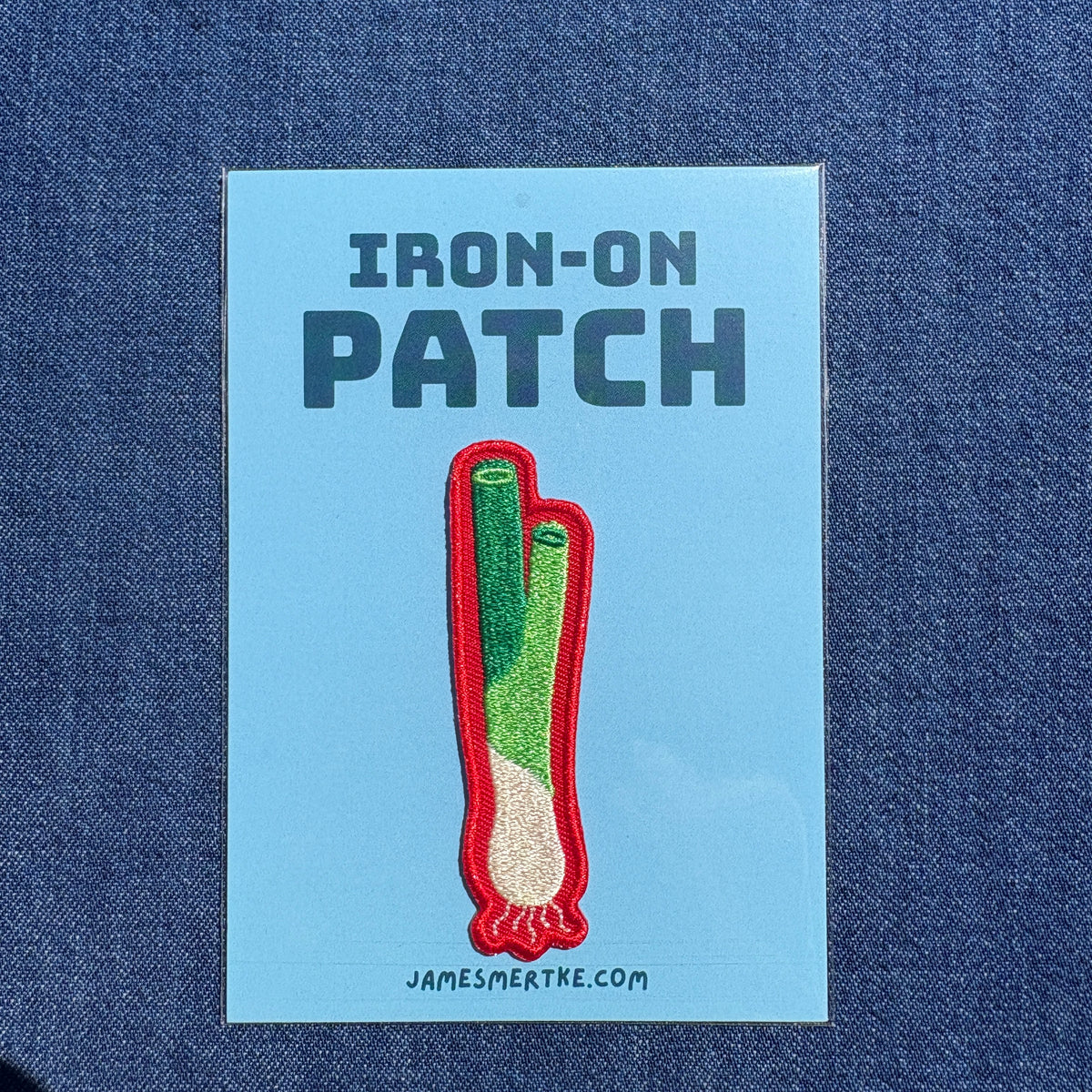 An iron-on patch featuring a green onion with a red border, presented on a light blue backing card that reads 'IRON-ON PATCH' and 'JAMESMERTKE.COM', set against a dark blue denim fabric background.