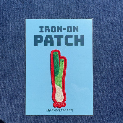 An iron-on patch featuring a green onion with a red border, presented on a light blue backing card that reads 'IRON-ON PATCH' and 'JAMESMERTKE.COM', set against a dark blue denim fabric background.