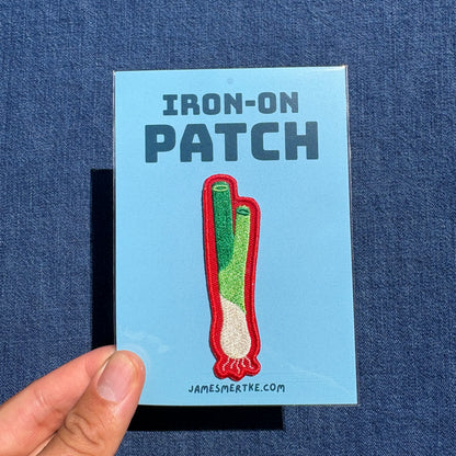 A hand holds a light blue backing card featuring an embroidered patch design of a green onion with the text 'IRON-ON PATCH' and 'JAMESMERTKE.COM'.