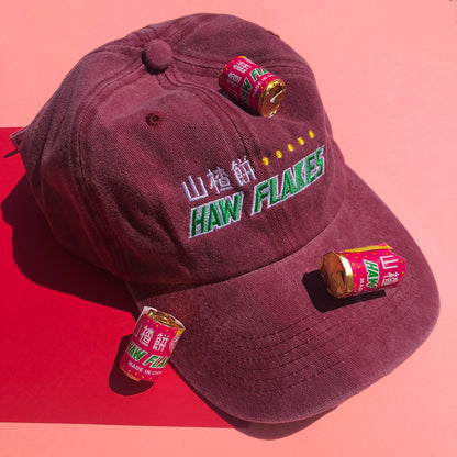 A dark red dad hat is embroidered with a design based off the haw flake candy. "Hawflakes" and its Chinese characters are written in white and green thread. Haw flake candies are scattered around the hat as props.