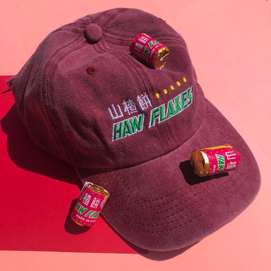 A dark red dad hat is embroidered with a design based off the haw flake candy. "Hawflakes" and its Chinese characters are written in white and green thread. Haw flake candies are scattered around the hat as props.