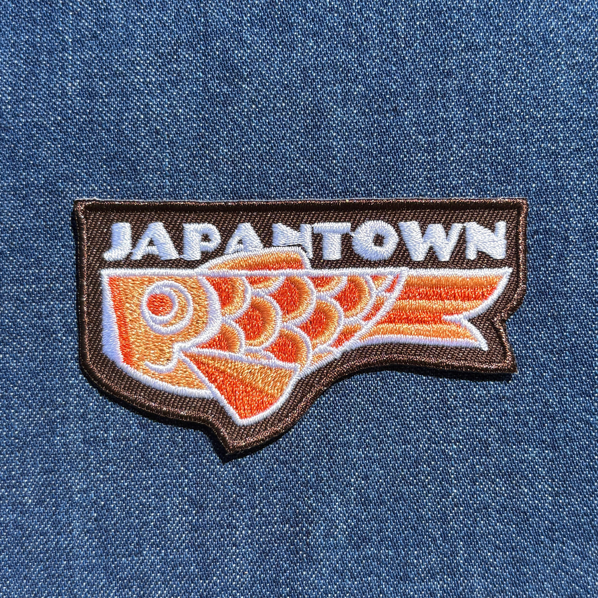 An embroidered patch featuring the word 'JAPANTOWN' in white above a stylized orange and white koinobori carp fish on a brown outline, shown on a blue denim background.