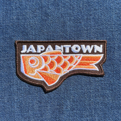 An embroidered patch featuring the word 'JAPANTOWN' in white above a stylized orange and white koinobori carp fish on a brown outline, shown on a blue denim background.
