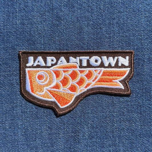 An embroidered patch featuring the word 'JAPANTOWN' in white above a stylized orange and white koinobori carp fish on a brown outline, shown on a blue denim background.