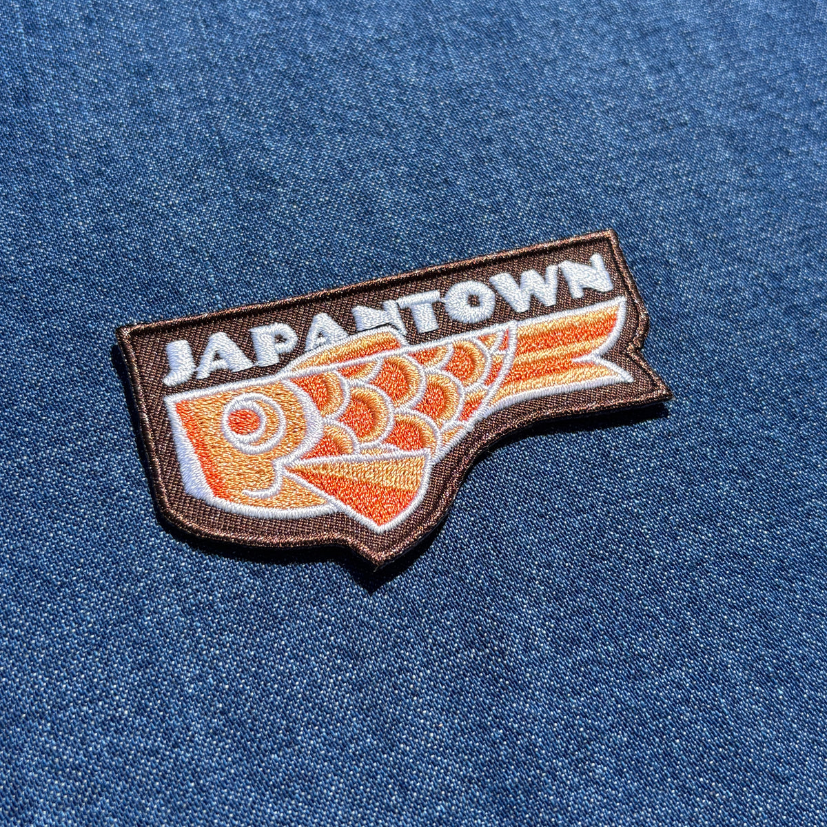 An embroidered patch featuring the word 'JAPANTOWN' in white above an orange and white stylized fish design, outlined in brown and placed on a blue denim fabric background.
