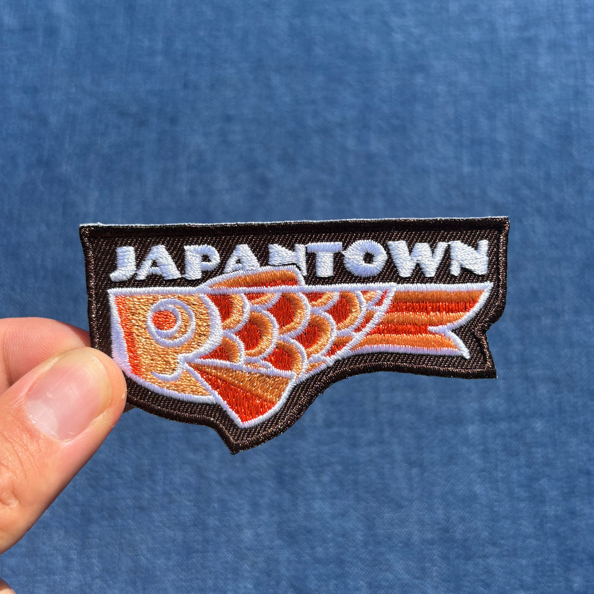 A close-up of a hand holding an embroidered patch that reads 'JAPANTOWN' above a stylized orange and white fish on a brown background, set against a blue textured fabric backdrop.