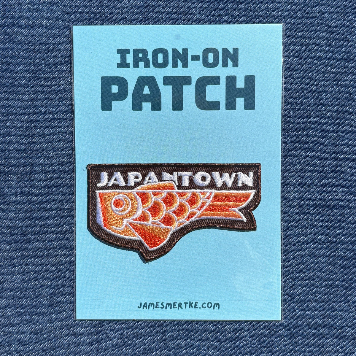 An iron-on patch displayed on a light blue backing card that reads 'IRON-ON PATCH' at the top and 'JAPANTOWN' above an orange and white fish graphic with a brown border. The card is placed on a blue denim background, with 'JAMESMERTKE.COM.