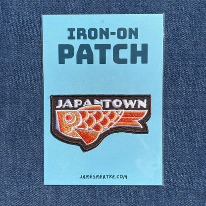 An iron-on patch displayed on a light blue backing card that reads 'IRON-ON PATCH' at the top and 'JAPANTOWN' above an orange and white fish graphic with a brown border. The card is placed on a blue denim background, with 'JAMESMERTKE.COM.