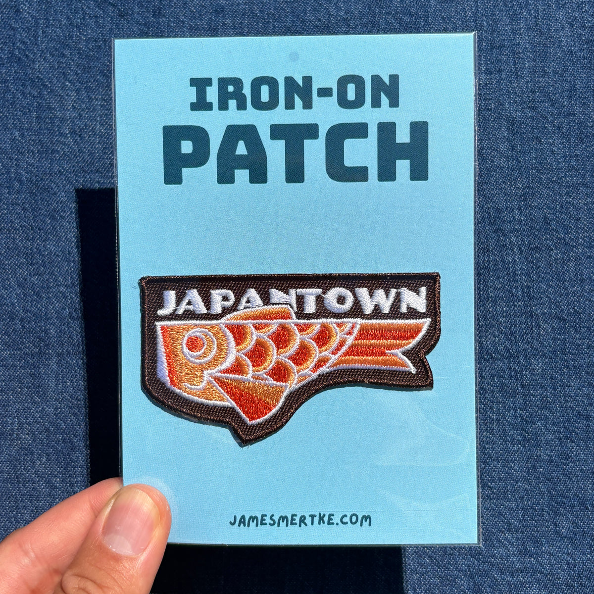 An Japantown fish iron-on patch displayed on a light blue backing card held by a hand against a dark blue denim background. The backing card reads 'IRON-ON PATCH' at the top and 'JAMESMERTKE.