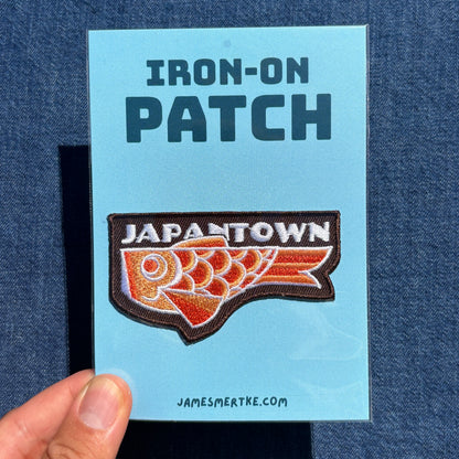 An Japantown fish iron-on patch displayed on a light blue backing card held by a hand against a dark blue denim background. The backing card reads 'IRON-ON PATCH' at the top and 'JAMESMERTKE.