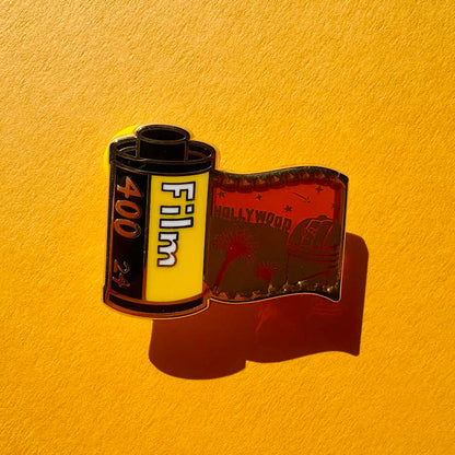 This pin features a film roll canister with "Film 400 24" written on it. It features a "stained glass" transparent panel with a screen-printed detail of Los Angeles including the Hollywood sign, Griffith Observatory, and palm trees when held up to the light.