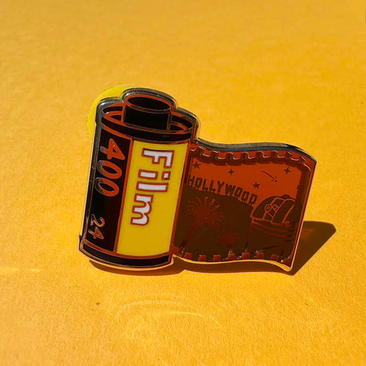 This pin features a film roll canister with "Film 400 24" written on it. It features a "stained glass" transparent panel with a screen-printed detail of Los Angeles including the Hollywood sign, Griffith Observatory, and palm trees when held up to the light.