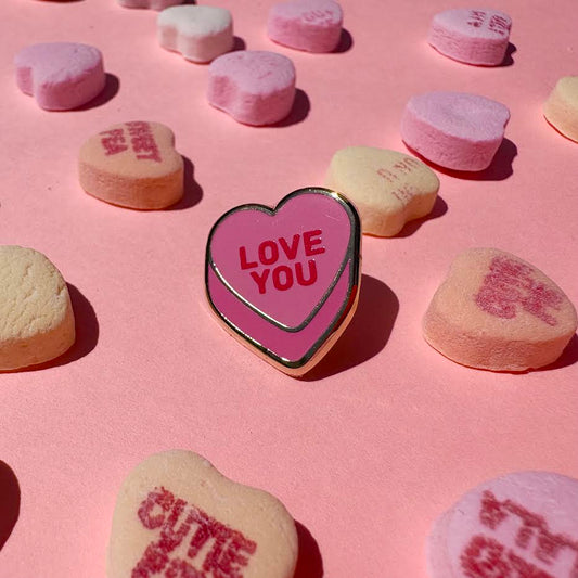 This enamel pin depicts a pink candy conversation heart with "LOVE YOU" written on the front in red text.

It is staged with candy hearts as props.