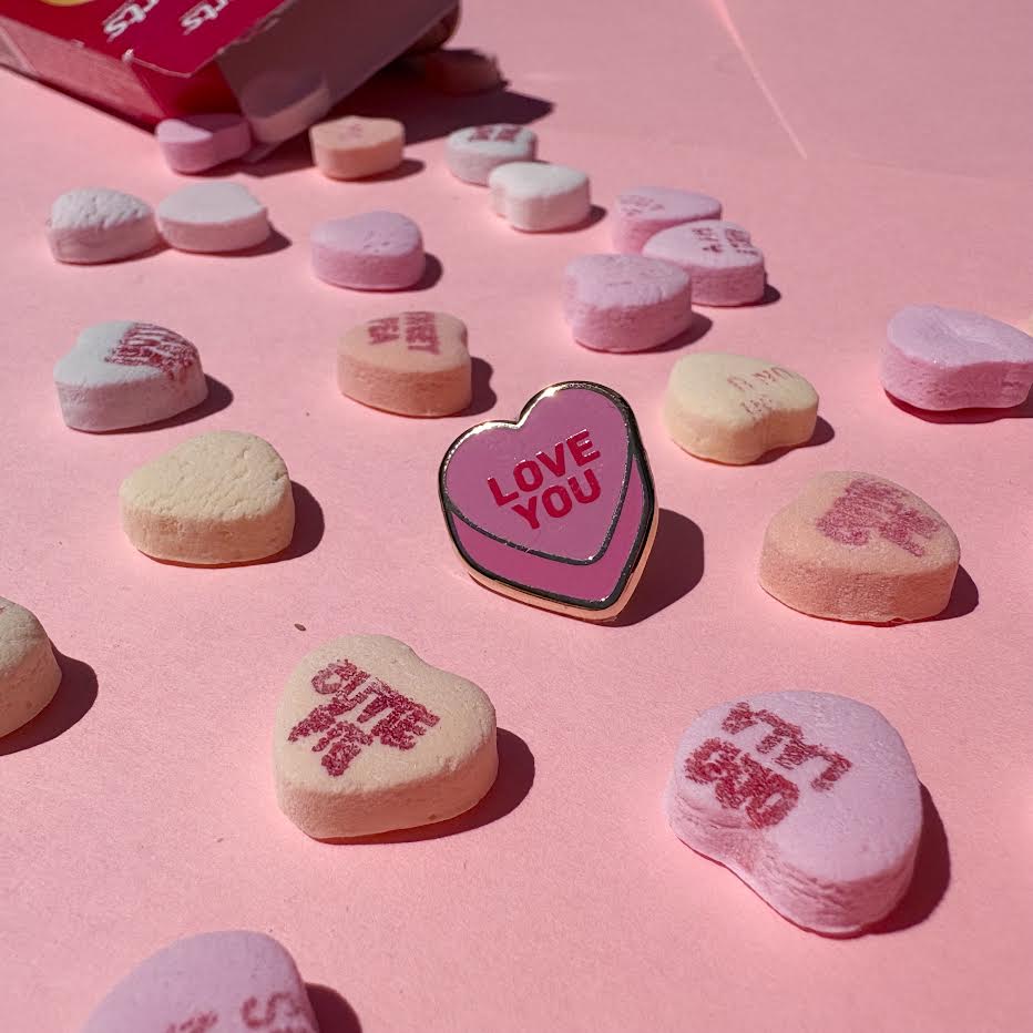 This enamel pin depicts a pink candy conversation heart with "LOVE YOU" written on the front in red text.

It is staged with candy hearts and a candy box as props.