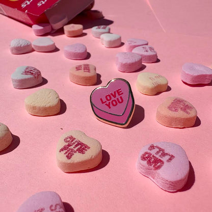This enamel pin depicts a pink candy conversation heart with "LOVE YOU" written on the front in red text.

It is staged with candy hearts and a candy box as props.