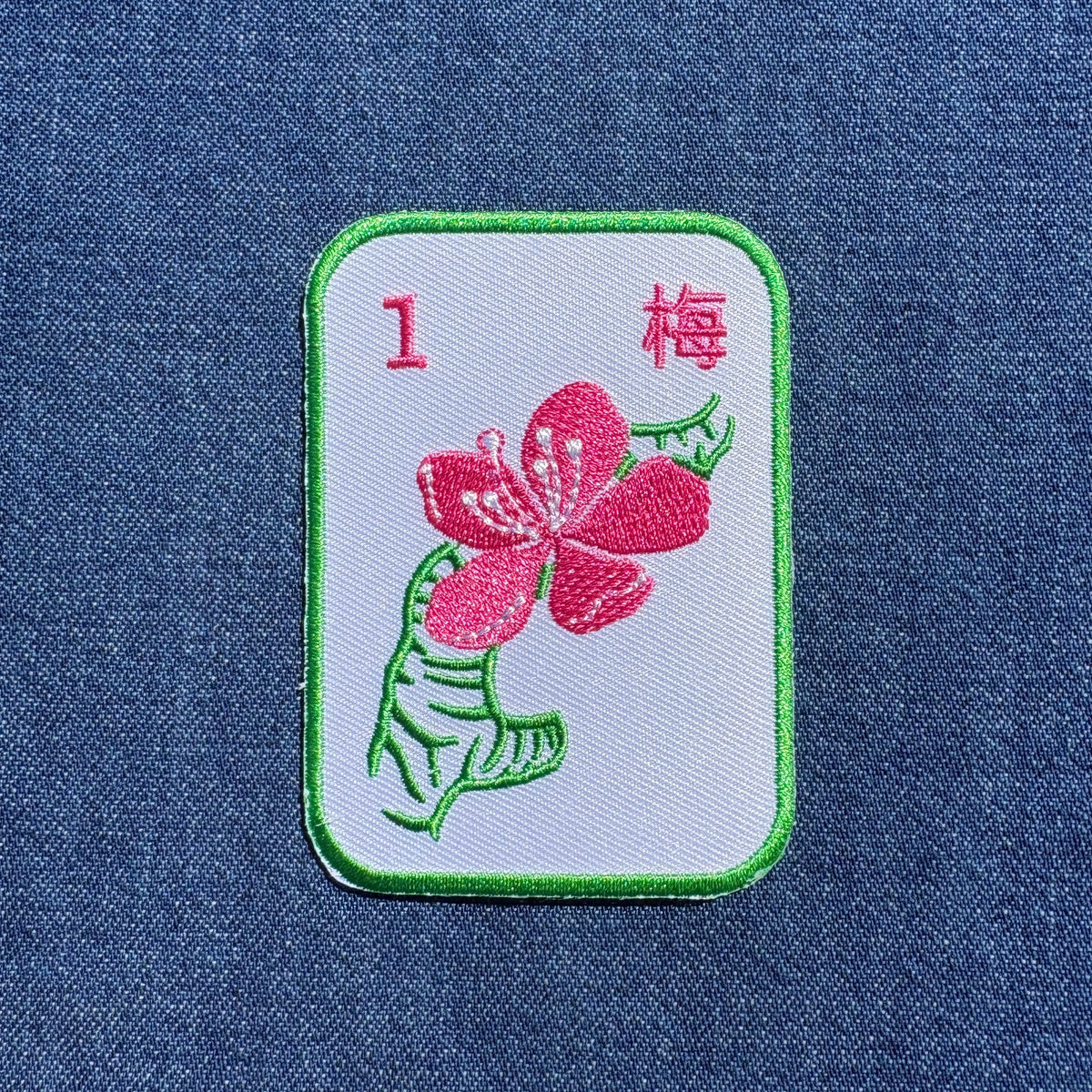 A rectangular embroidered patch depicting a mahjong tile pink plum blossom on a white background with green border, placed on a blue denim fabric.
