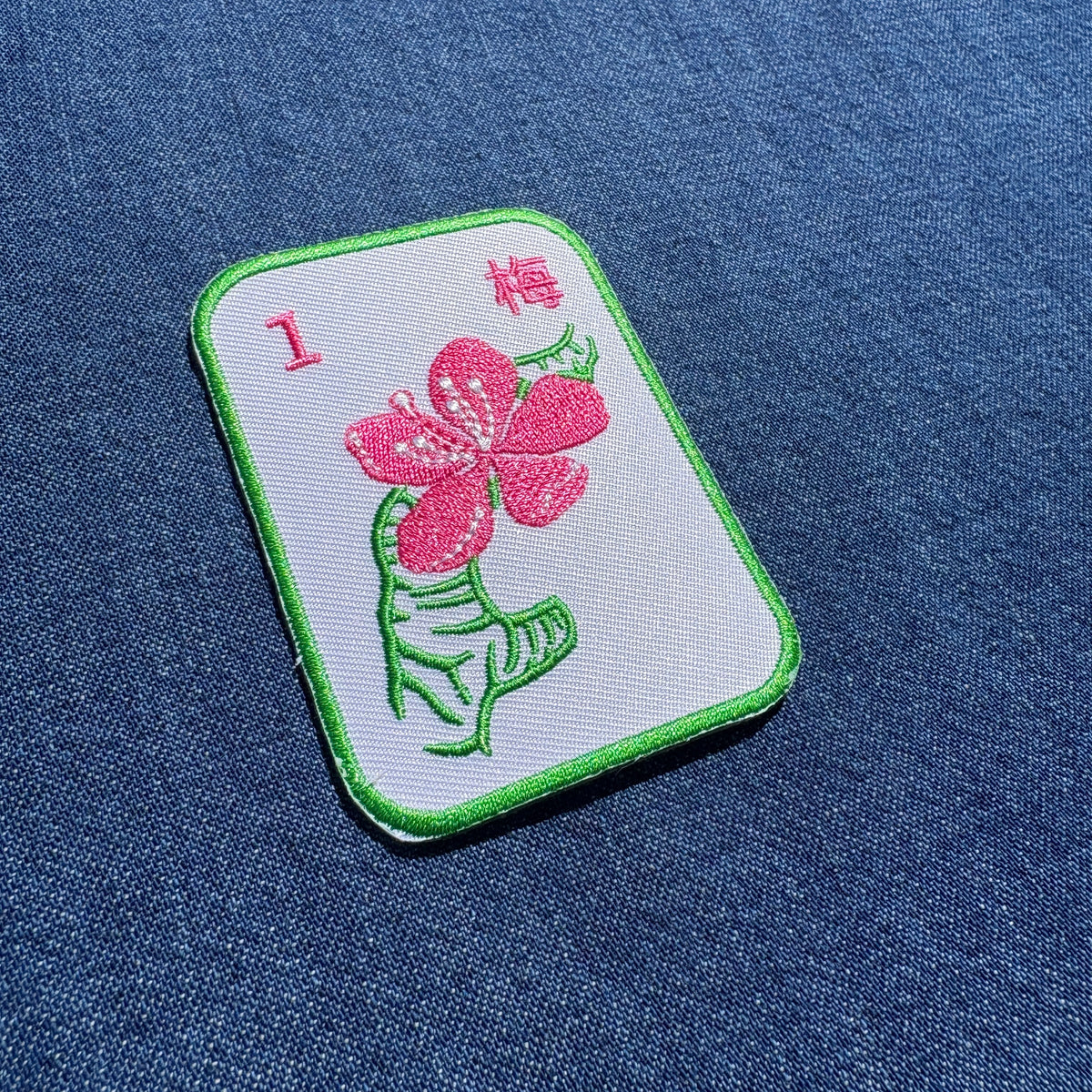 A rectangular embroidered patch with rounded corners, featuring a pink flower and green leaf motif on a white background with a green border, placed on a blue denim-like fabric surface.
