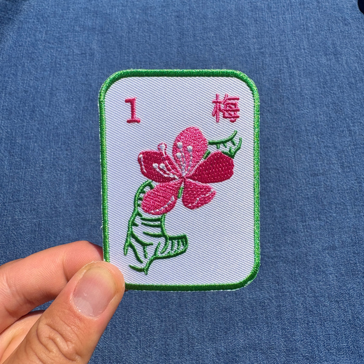 A hand holds a rectangular embroidered patch featuring a pink flower with green leaves on a white background, bordered in green. The patch includes the red number 1 and the Chinese character for plum.
