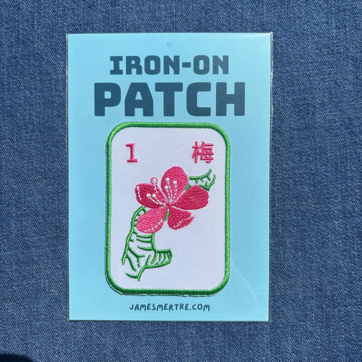 An iron-on patch displayed on a blue denim fabric background. The patch is on a light blue backing card with the text 'IRON-ON PATCH'. The patch features an embroidered design of a mahjong tile of a pink cherry blossom flower with green leaves on a white background, labeled with the number 1 and the Chinese character for plum.